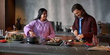 Woman in pink sweater cooking on hob next to man in red jacket cutting vegetables
