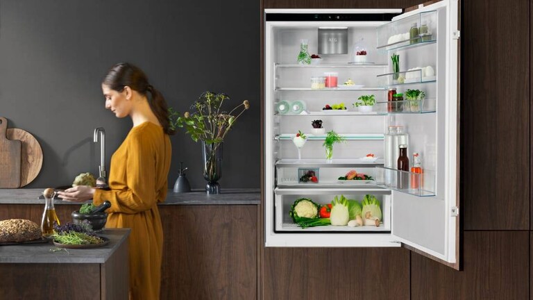 Nainen seisoo keittiössä avoinaisen jääkaapin edessä.